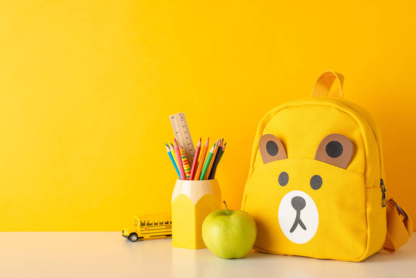 Навчання основам на виставці. Фотографія розташування столу, що демонструє кольорові олівці, лінійку, чарівний плюшевий ведмедик рюкзак, шкільну автобусну іграшку та зелене яблуко на жовтому тлі стіни - Фото, зображення