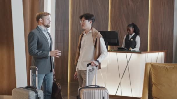 Young adult Asian and Caucasian men on business trip standing in hotel lobby with their suitcases discussing something - Footage, Video