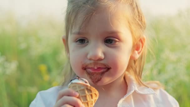 Lähikuva valkoihoinen tyttö syö jäätelöä nurmikolla kentällä. Onnellinen hymyilevä lapsi peitetty kasvovoide syöminen jälkiruoka. Laadukas 4k kuvamateriaalia - Materiaali, video