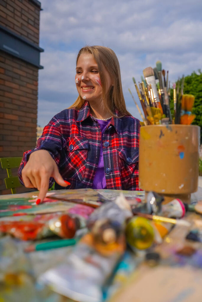 Retrato de una artista alegre pintando con el dedo pintura en su cara sentada en una mesa de pintura con tubos de pintura y pinceles en ella - Foto, Imagen