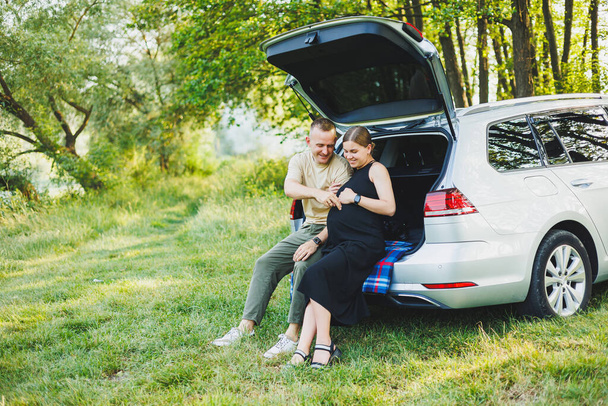 Счастливая молодая супружеская пара, ожидающая ребенка, сидит в багажнике автомобиля в природе. Мужчина и женщина обнимают беременный живот, сидя на зеленой лужайке - Фото, изображение