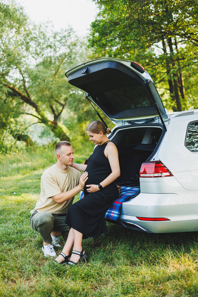 Ein glückliches junges Ehepaar, das ein Kind erwartet, sitzt im Kofferraum eines Autos in der Natur. Ein Mann und eine Frau umarmen einen schwangeren Bauch, während sie auf einem grünen Rasen sitzen - Foto, Bild