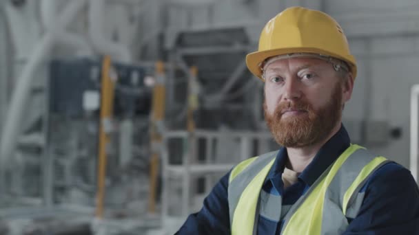 Medium close-up slow motion van knappe volwassen blanke man met baard op gezicht met gele hardhoed werkend in fabriek kijkend naar camera - Video