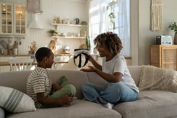 Young African American mother holding VR helmet introducing virtual reality technology to child son while spending leisure time together at home. Mom parent exploring virtual world with kid - Fotó, kép