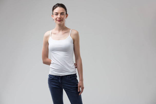 beautiful, slim woman exudes a hint of androgyny or tomboyish charm. With her hair pulled back, wearing a white tank top and blue jeans, she coyly keeps an arm behind her back, subtly showcasing her e - Photo, Image