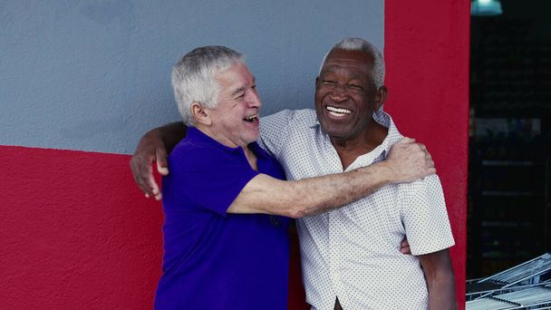 Autentyczne interakcje dwóch szczęśliwych, zróżnicowanych, starszych przyjaciół, przytulanie i świętowanie z przybitą piątką, stojących na zewnątrz na chodniku. Wesołe towarzystwo pomiędzy Afroamerykanami a osobami rasy kaukaskiej - Zdjęcie, obraz