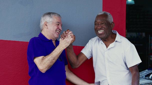 Authentic Interaction of Two Happy Diverse Older Friends, Hugging and Celebrating with High-Five, Standing Outside on Sidewalk. Cheerful Companionship Between African American and Caucasian Individuals - Photo, Image