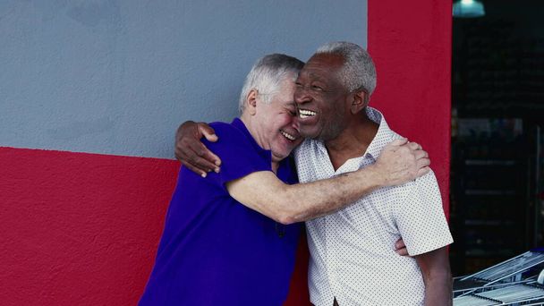 Authentische Interaktion zweier glücklicher älterer Freunde, die sich umarmen und mit High-Five feiern, draußen auf dem Gehweg stehen. Fröhliche Kameradschaft zwischen Afroamerikanern und Kaukasiern - Foto, Bild