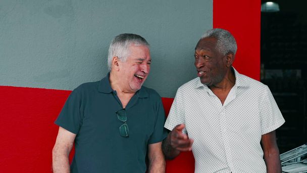 Radosna interakcja dwóch różnych męskich starszych przyjaciół, uśmiechniętych i śmiejących się razem, pochylonych na ścianie chodnika. Autentyczne prawdziwe życie szczęśliwych starszych ludzi - Zdjęcie, obraz