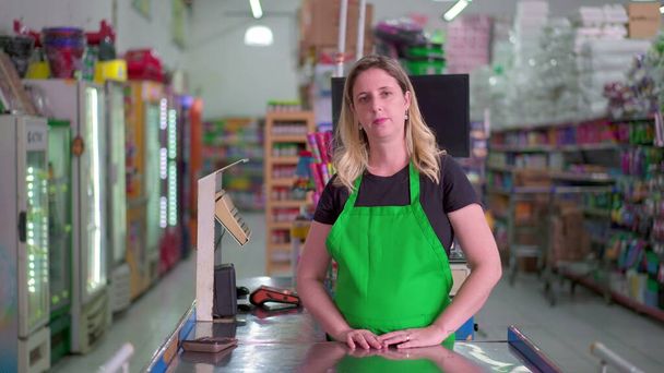 Kobieta pracownik supermarketu stoi przy kasie i patrzy w kamerę z neutralnym wyrazem twarzy w zielonym fartuchu. Portret personelu Sklepu Spożywczego - Zdjęcie, obraz