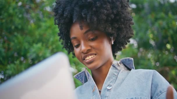 Zwart-harige vrouw videocalling door laptop zittend onder groene bomen close-up. Portret van een prachtig glimlachend Afrikaans Amerikaans meisje dat praat met een computer webcamera buiten. Leuke freelancer die online spreekt - Video