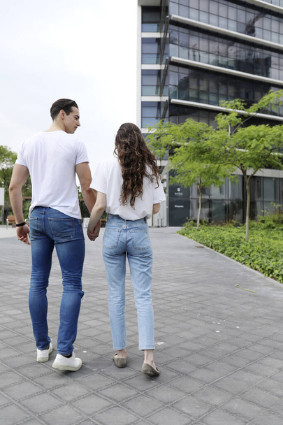 Rear view of unrecognizable young couple in love wearing casual clothes walking together on city street and holding hands. Loving man and woman enjoying leisure time together outdoors on summer day. - Foto, Bild