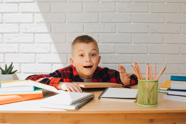 Концепция вернуться в школу. Веселый мальчик в процессе учебы сидит за столом с книгами, блокнотами и карандашами. Вид спереди - Фото, изображение
