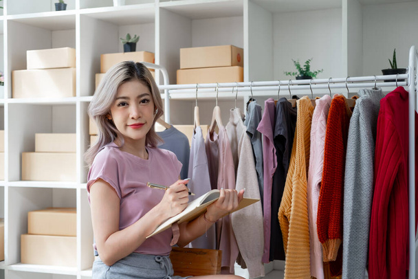 Young woman female owner business online selling checking clothes online and hold book and pen for writing - 写真・画像