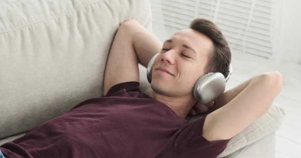 Un homme caucasien est capturé allongé sur le canapé dans le salon, portant un sourire de contentement alors qu'il écoute de la musique. Son visage respire le bonheur et le plaisir. - Séquence, vidéo