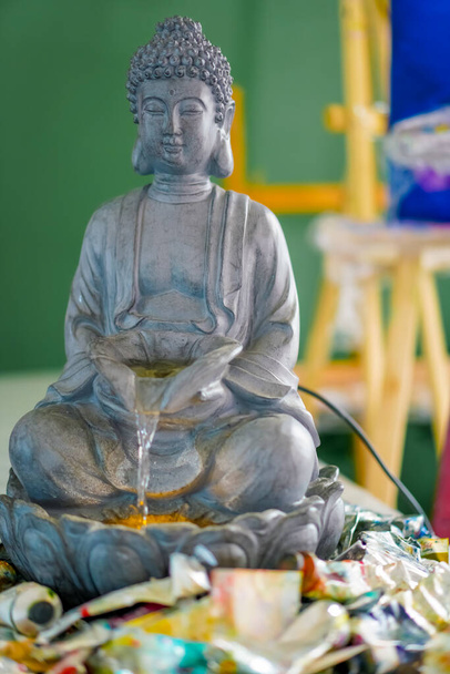Um close-up da estátua da fonte de Buda em pé sobre uma mesa em um estúdio de pintura com tubos de tinta ao lado dele - Foto, Imagem