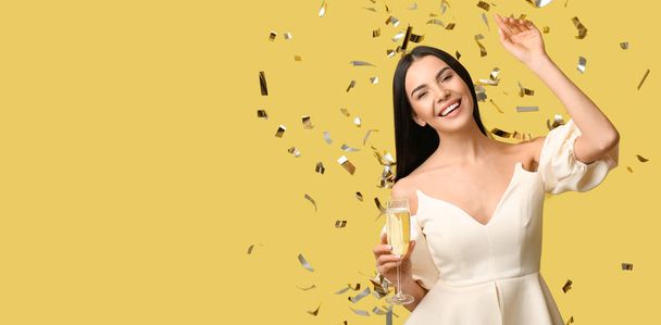 Belle femme heureuse avec verre de champagne et confettis tombant sur fond jaune avec espace pour le texte - Photo, image