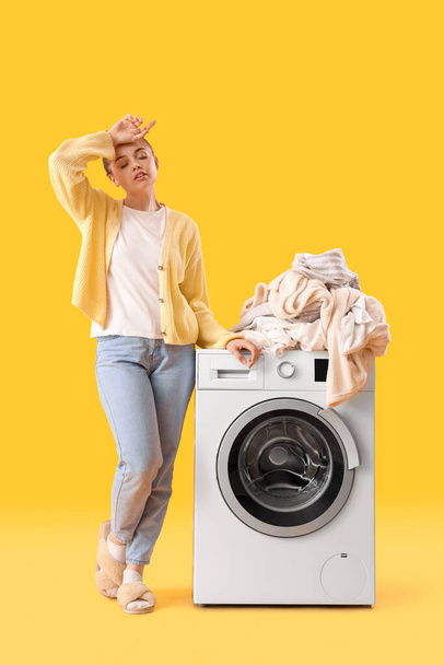 Κουρασμένη νεαρή γυναίκα στέκεται κοντά στο πλυντήριο με σωρό από βρώμικα ρούχα σε κίτρινο φόντο - Φωτογραφία, εικόνα