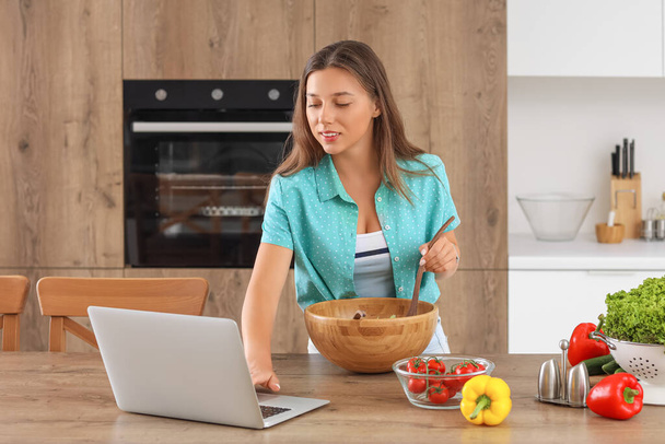 Jonge vrouw met laptop het bereiden van groentesalade in de keuken - Foto, afbeelding