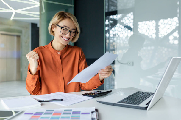 Young happy and successful female financier inside the office at the workplace is satisfied with the results of the achievement, smiling and reading report contract, businesswoman at work paperwork. - Foto, Imagem