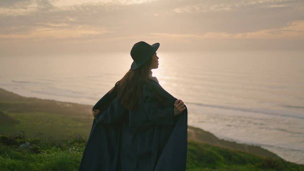 Силуэт молодой девушки, позирующей на живописном осеннем закате. Серьёзная задумчивая женщина, стоящая перед закатом над прекрасным океаном. Модель с длинными волосами в стильном пальто в шляпе наслаждается вечером на природе - Фото, изображение
