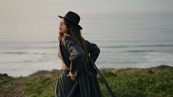 Stylish gorgeous girl looking sunset standing alone on green hill near calm ocean. Young woman wearing elegant coat hat watching amazing sea sundown at autumn evening. Relaxed model posing on camera. - Photo, image