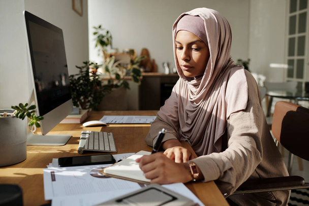 Νεαρή σοβαρή μουσουλμάνα επιχειρηματίας κρατώντας σημειώσεις στο σημειωματάριο ή έγγραφο, ενώ κάθεται στο χώρο εργασίας μπροστά από την οθόνη του υπολογιστή - Φωτογραφία, εικόνα