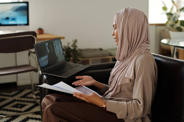 Nuori itsevarma musliminainen, jolla on taloudellisia papereita, katselee kollegaa kannettavan tietokoneen näytöllä kommunikoidessaan videokeskustelussa digitaalisessa kokouksessa - Valokuva, kuva