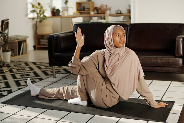 Mladá aktivní žena v hidžábu a kombinéze sedí na podložce na podlaze obývacího pokoje a drží pravou dlaň otevřenou během cvičení jógy - Fotografie, Obrázek