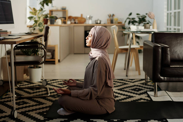 Nuori rauhallinen nainen hijabissa ja haalareissa istuu lootuksen asennossa matolla työpaikan edessä tietokonenäytön ja meditoinnin kanssa - Valokuva, kuva