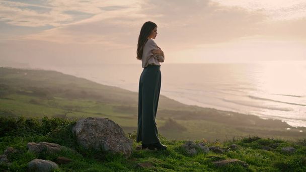 若い魅力的な女性が夕日と素晴らしい景色を見て緑の芝生の丘に立っている。リラックスしたロングヘアのパッシブモデルが海岸をポーズ。女の子交差手上のボディ身に着けているスタイリッシュなカジュアル服 - 写真・画像