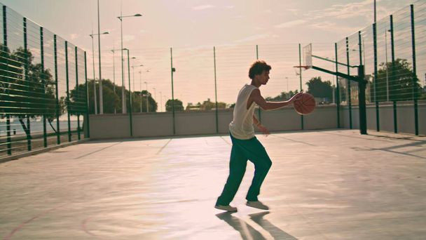 Involved teenager bounce ball at stadium. Basketball player making basket throw at urban playground. Curly young man training court. Sportive guy playing summer morning. Workout and sport activities - Foto, Bild
