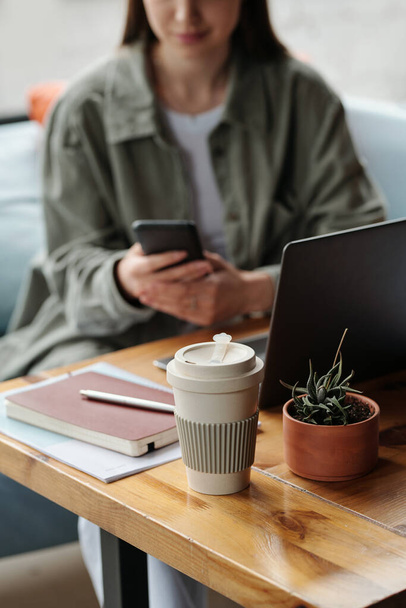 Concentre-se em vidro descartável de café e vaso de flores marrom com planta verde no local de trabalho do gerente feminino usando smartphone na frente do laptop - Foto, Imagem