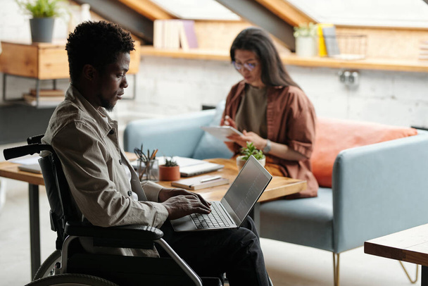 Visão lateral do jovem afro-americano sério buinessman em cadeira de rodas digitando no teclado do laptop enquanto trabalhava sobre novo projeto no escritório - Foto, Imagem