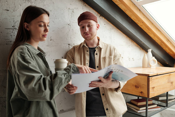 Young confident businesswoman with cup of coffee pointing at paperwork with financial data while explaining it to male colleague at meeting - Foto, imagen