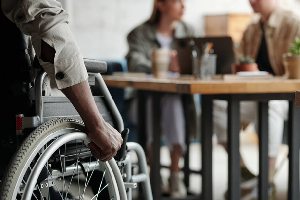 Część młodego Afroamerykanina męskiego pracownika z niepełnosprawnością trzymając rękę na kole wózka inwalidzkiego podczas ruchu w kierunku swoich kolegów - Zdjęcie, obraz