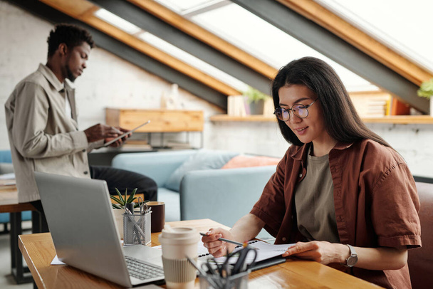 Fiatal mosolygós női elemző átnézi a pénzügyi dokumentumot, miközben ül a munkahelyen előtt laptop ellen férfi kolléga - Fotó, kép