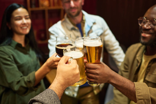 Focus op handen van vier jonge interculturele vrienden klinkend met een glas fris koud bier terwijl ze plezier hebben en genieten van feest of bijeenkomst in de bar - Foto, afbeelding