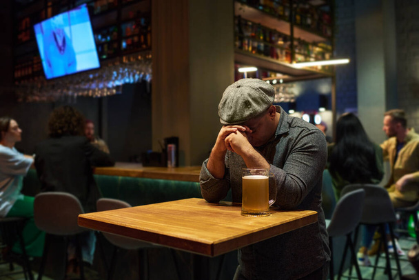 Joven hombre deprimido con las manos a la cara inclinándose sobre la mesa con taza de cerveza en el bar mientras sufre de dolor o se siente apenado o ansioso - Foto, Imagen