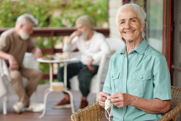 Веселая пожилая женщина с белыми волосами, смотрящая в камеру, сидя в плетеном кресле на террасе дома престарелых и вязав шарф - Фото, изображение