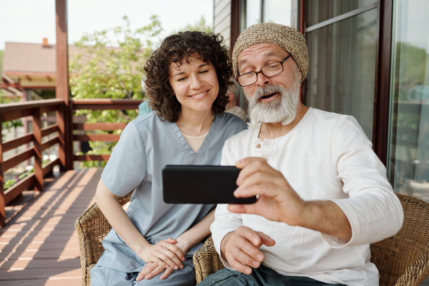 Jeune soignant heureux et homme âgé regardant un film ou une vidéo en ligne sur smartphone assis dans des fauteuils sur le patio de la maison de retraite - Photo, image