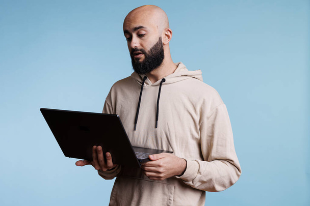 Hombre árabe serio sosteniendo portátil y participando en videoconferencia en línea. Joven calvo usando computadora portátil para conversación remota usando software de videocall web - Foto, imagen