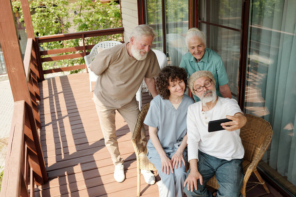Gelukkig bejaarden en hun verzorger selfie nemen op patio van bejaardentehuis, terwijl een van hen houden smartphone in de voorkant van hun gezicht - Foto, afbeelding