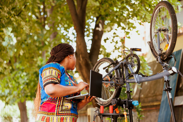 Zdravý a živý afroameričtí fermale výzkum na internetu pro údržbu jízdních kol pro venkovní volný čas cyklistiky. Sportovně-milující černošky žena kontrolu na kole zařízení na notebooku. - Fotografie, Obrázek