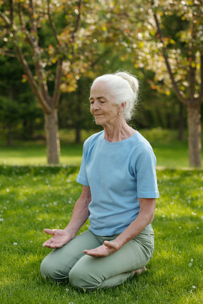 Uśmiechnięta starsza kobieta z siwymi włosami siedząca w pozycji lotosu na zielonej trawie i ćwicząca jogę podczas terapii rehabilitacyjnej - Zdjęcie, obraz
