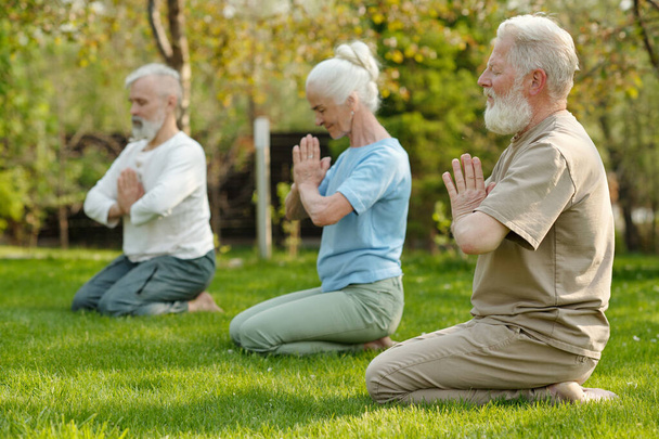 Concéntrese en el hombre mayor barbudo manteniendo las manos juntas por el pecho mientras practica ejercicio de yoga con otros pacientes de la casa de retiro - Foto, Imagen