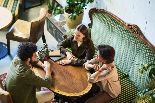 Під кутом трьох молодих міжкультурних друзів спілкуються, сидячи за круглим дерев'яним столом у кафе або ресторані з старовинними меблями - Фото, зображення