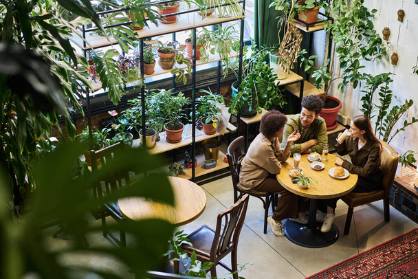Dwóch międzykulturowych przyjaciół lub partnerów rozmawiających w kawiarni, siedzących przy stole obok młodej kobiety pijącej latte i słuchających ich dyskusji - Zdjęcie, obraz