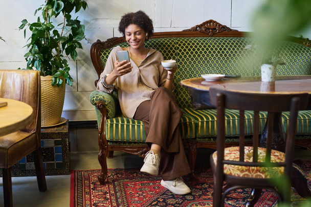 Ευτυχισμένη νεαρή ξεκούραστη Αφροαμερικανή γυναίκα που τρέχει στο smartphone ή βλέπει online βίντεο και πίνει καφέ ενώ κάθεται στον καναπέ - Φωτογραφία, εικόνα