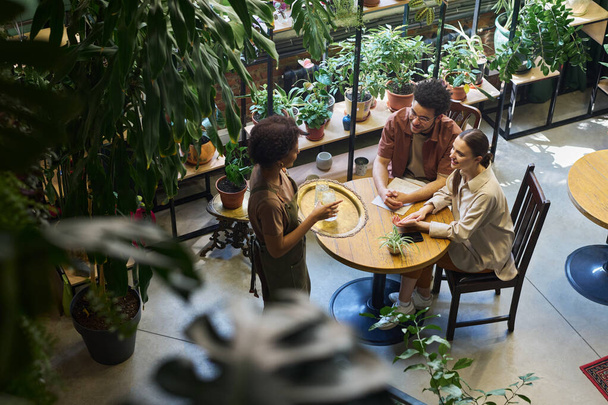 Високий кут молодої міжкультурної пари, дивлячись на офіціантку з підносом, що стоїть перед їх столом в кафе, приносячи їм лимонад - Фото, зображення